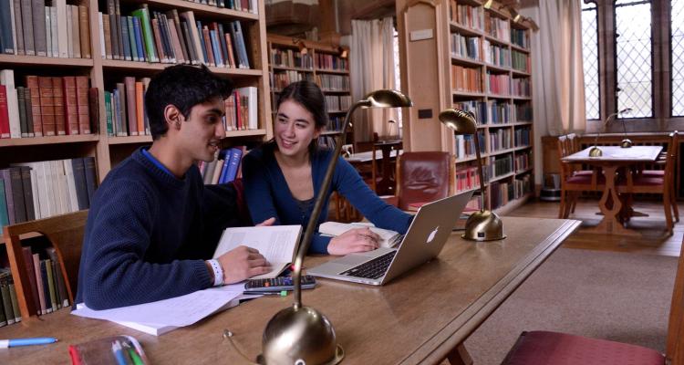 At a table between bookshelves, two students discuss their work over a laptop and papers. Warm light streams form a window, warming the reds, browns and blues of the books and bookshelves.  